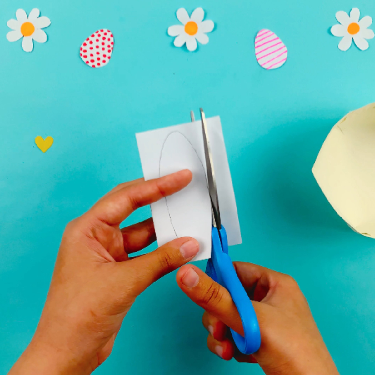 Two hands taking folded piece of rectangular white paper and cutting out the shape of bunny ears that have been drawn on with a pencil.