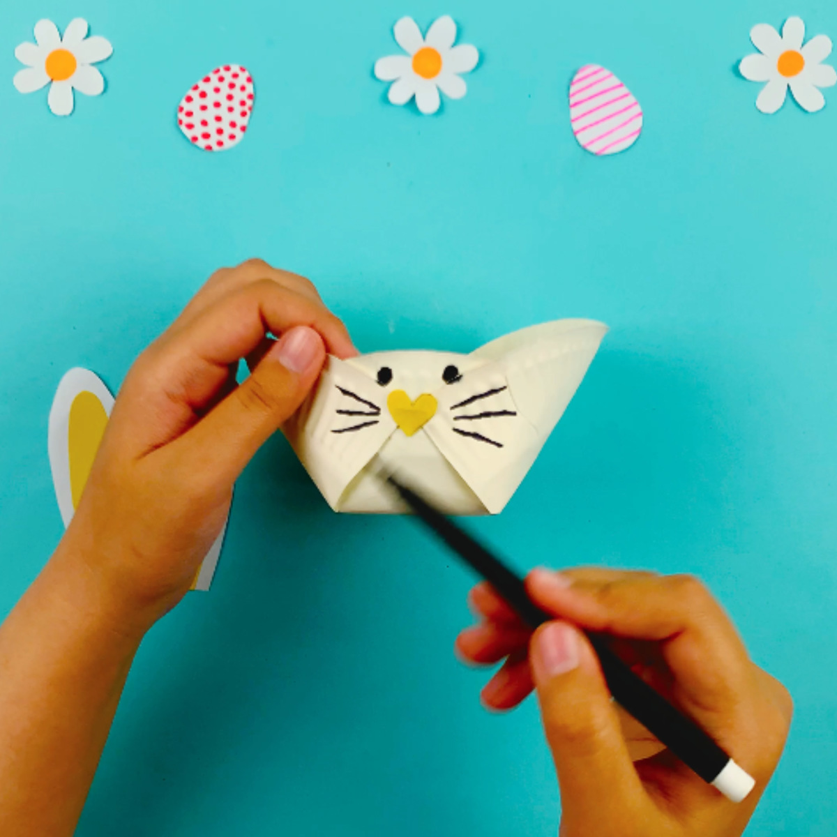 Two hands drawing a bunny face on the paper plate basket after the nose has been stuck on.