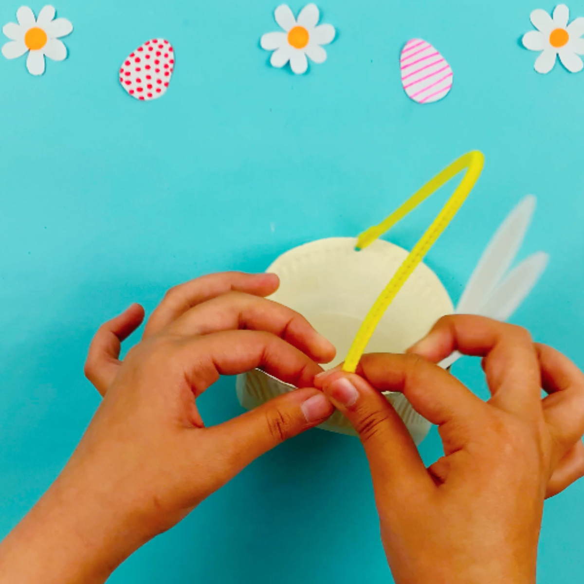Two hands using a punch to make holes in the side of the paper plate basket and then using a pipe cleaner to form the handle.
