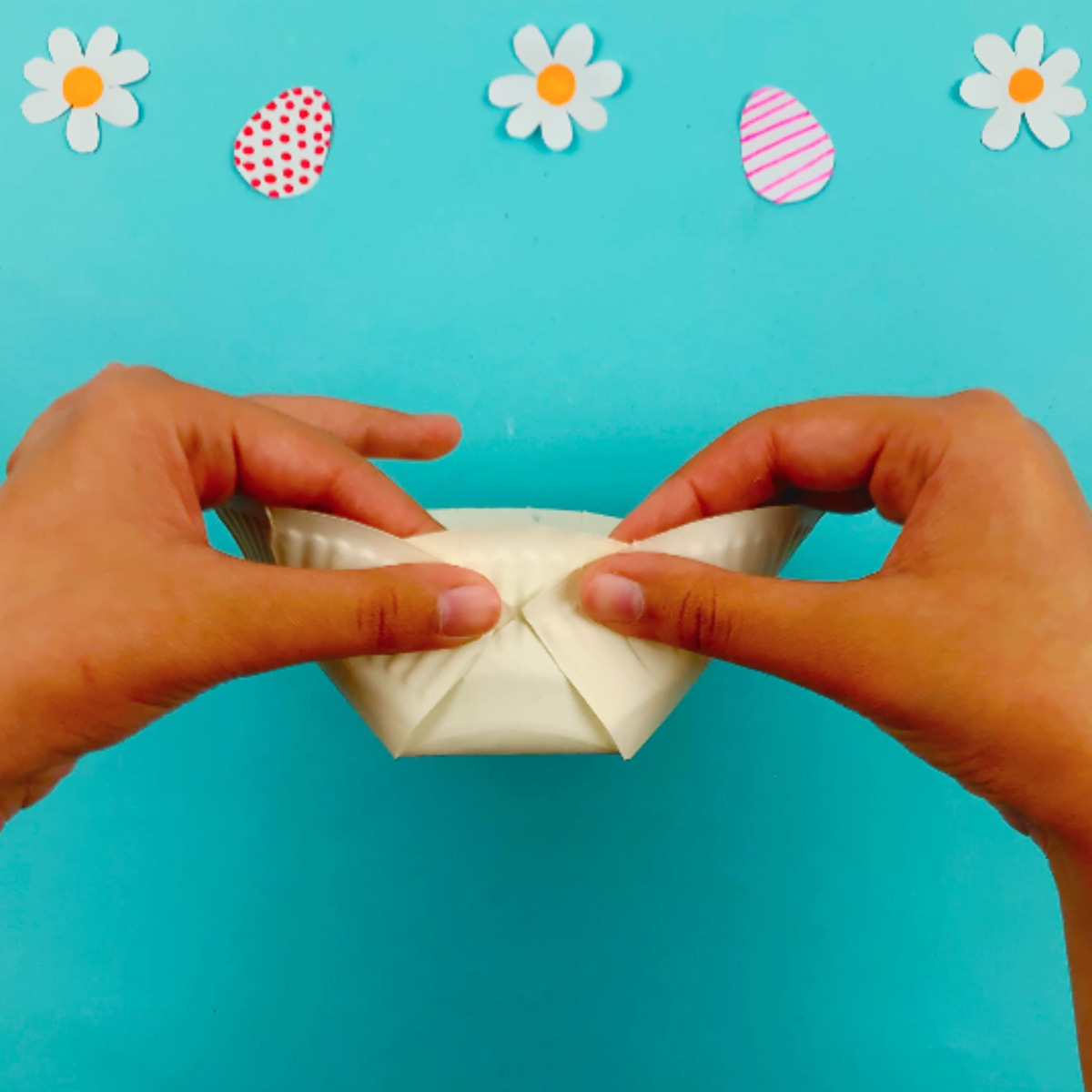 Two hands folding the cut sections on the bottom of the plate, both the left and right hand side and attaching them to the middle with a staple.
