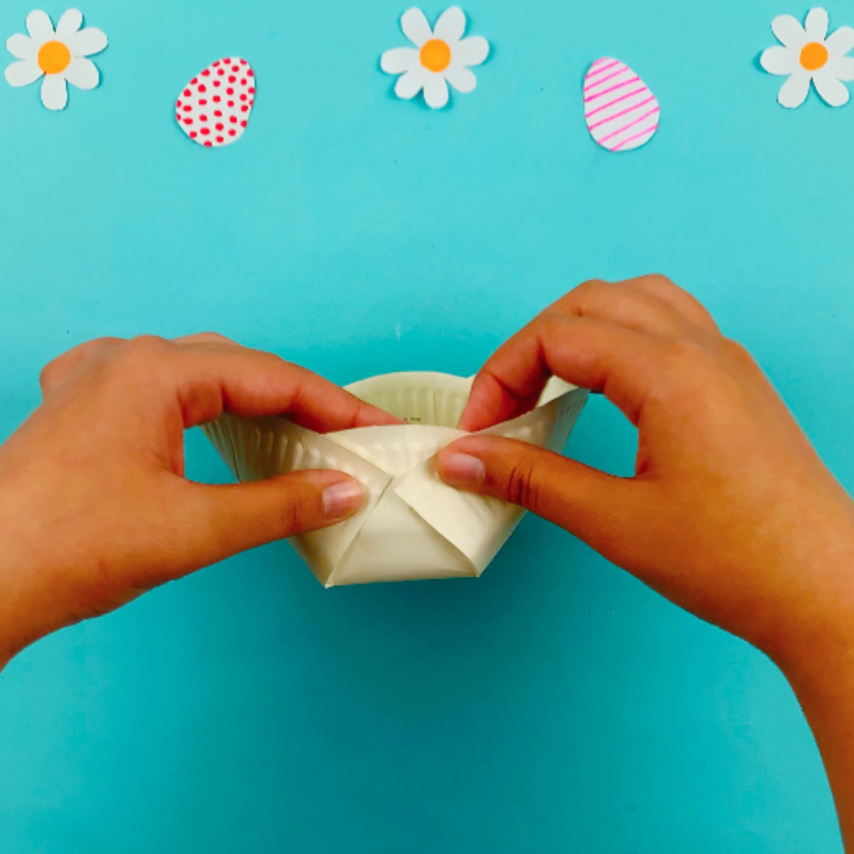 Two hands folding the top section of the plate, both the left and right hand sides to the middle section and attaching them with a staple to form a basket shape.