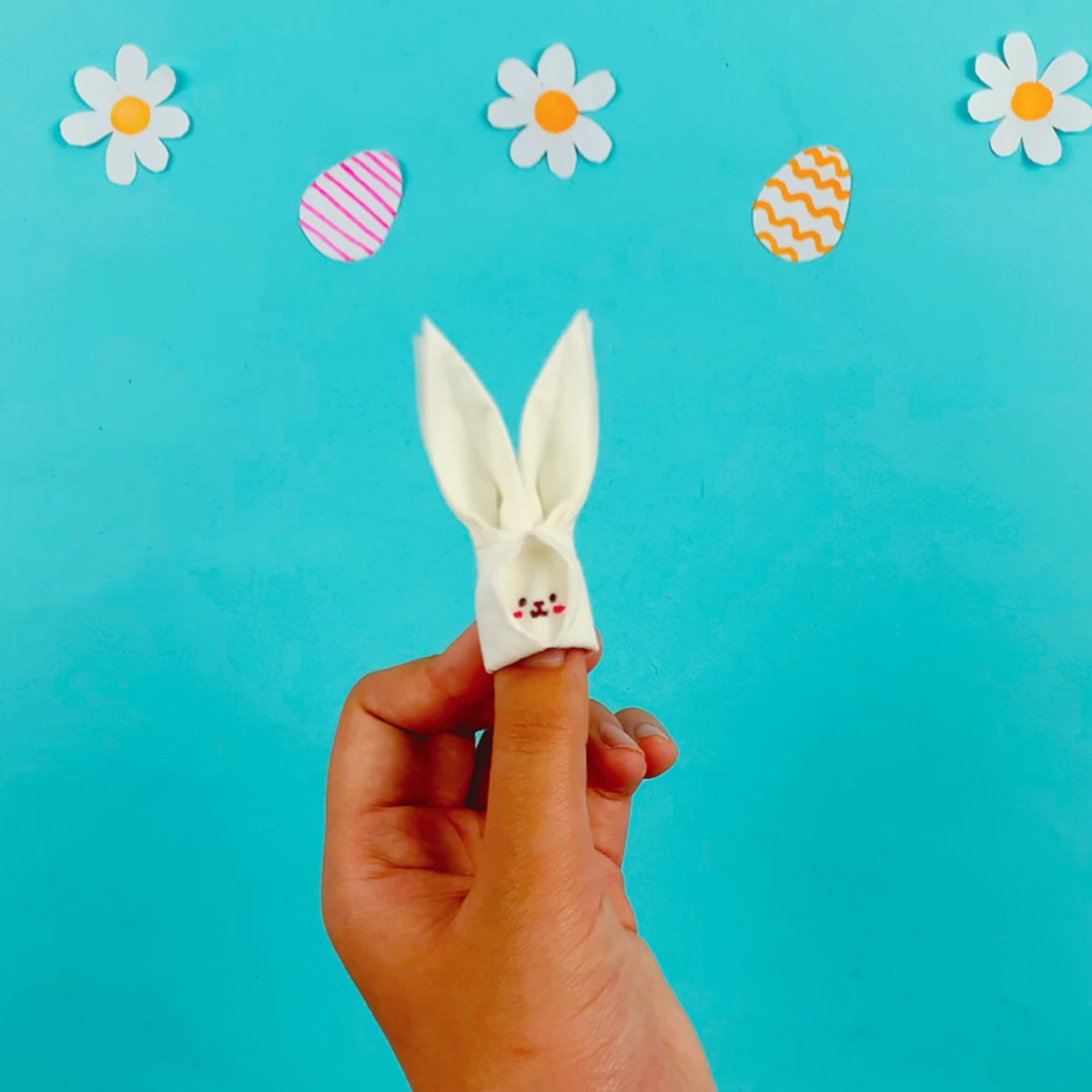 Two hands holding the tissue bunny finger puppet after drawing on the face using colored pens.