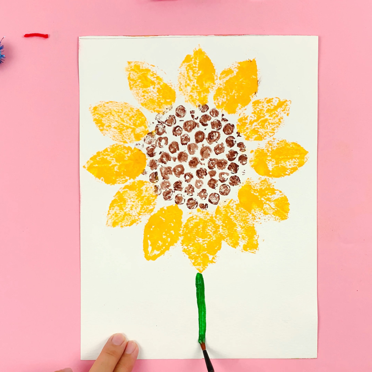 A hand showing the stem painted downwards from the bottom sunflower petal using green paint.
