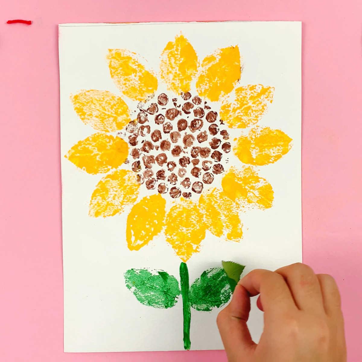 A hand using a painted garden leaf to stamp leaf shapes on the stem. 