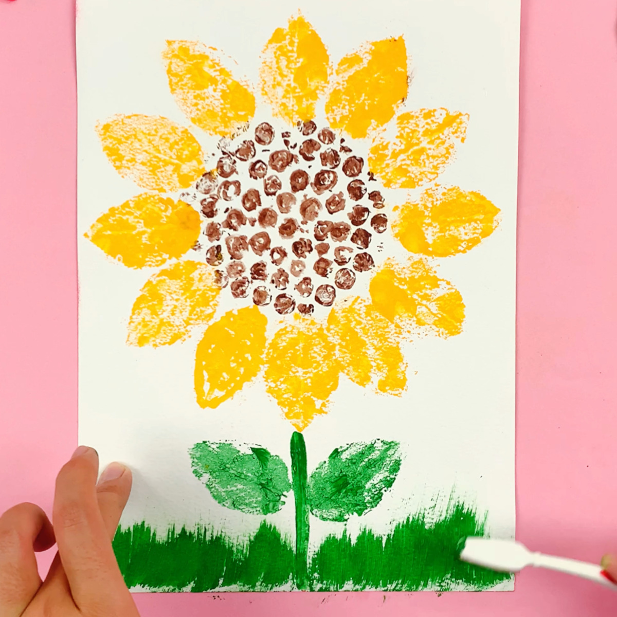 A hand using an old paintbrush with green paint added to paint grass at the bottom of the white cardstock.