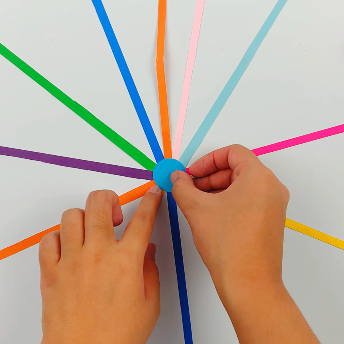 Two hands gluing a paper circle without a punched whole to secure all the colored strips of paper. 